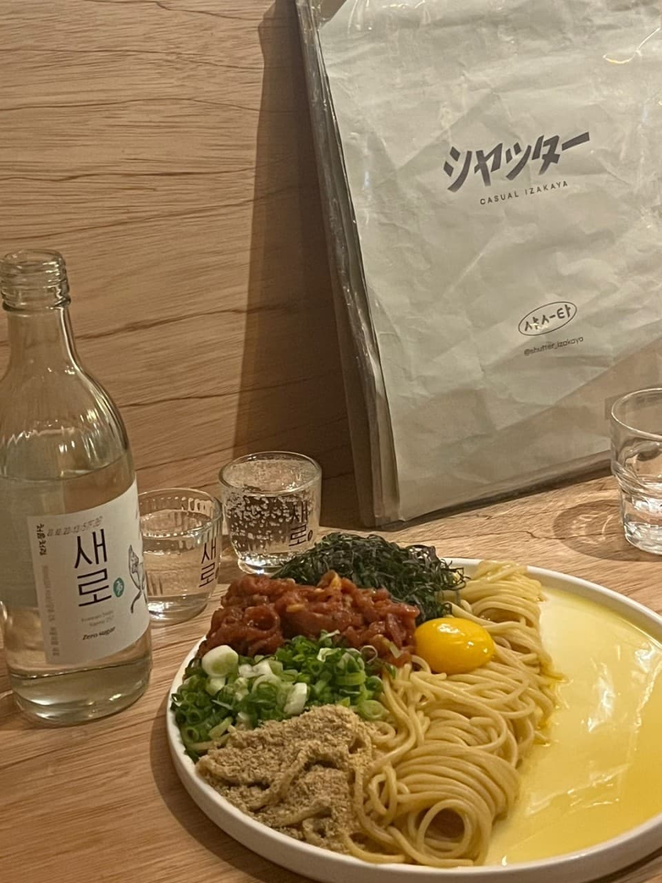 경상북도 경주시 혼술 이자카야 샷타 경북 - 나무 소재 다찌석 위 노란 조명이 비치는 노른자와 쪽파를 곁들인 비빔면과 새로 소주 한 병