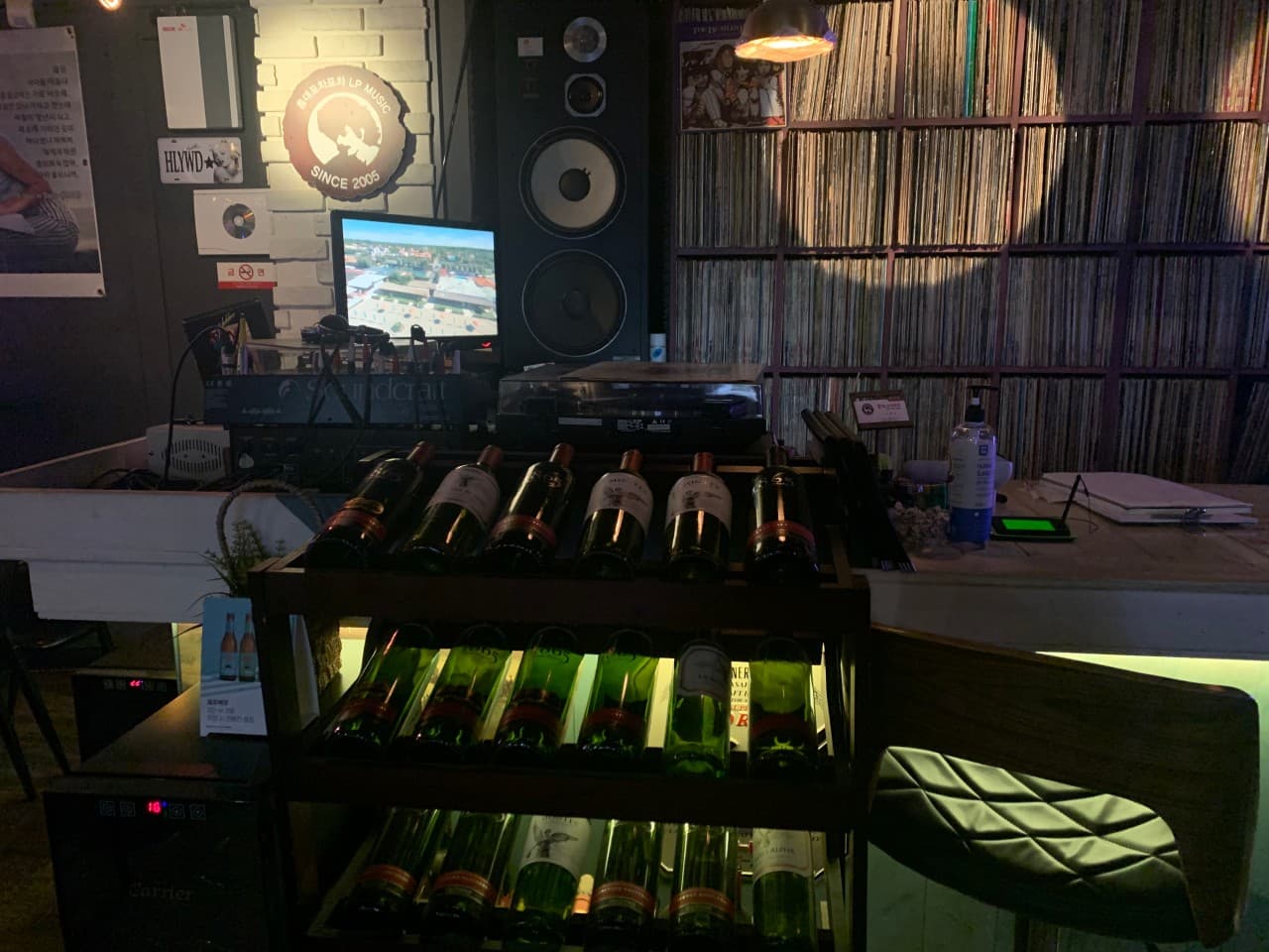 대전광역시 서구 홍대포차포차 대전의 LP 판으로 채워진 벽과 조명 아래 진열된 와인병들. 대형 스피커와 1인 다찌석이 마련된 공간에서 혼술을 즐기는 상황을 묘사함.