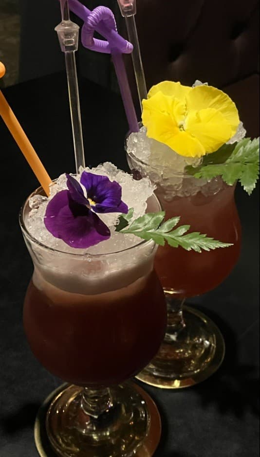 경상북도 경산시 바(Bar) 비사이드 경북의 어두운 테이블 위 보라색과 노란색 식용 꽃, 잘게 부순 얼음이 가득한 칵테일. 1인 혼술 상황에 어울리는 빨대가 꽂힌 음료의 질감을 보여줌.