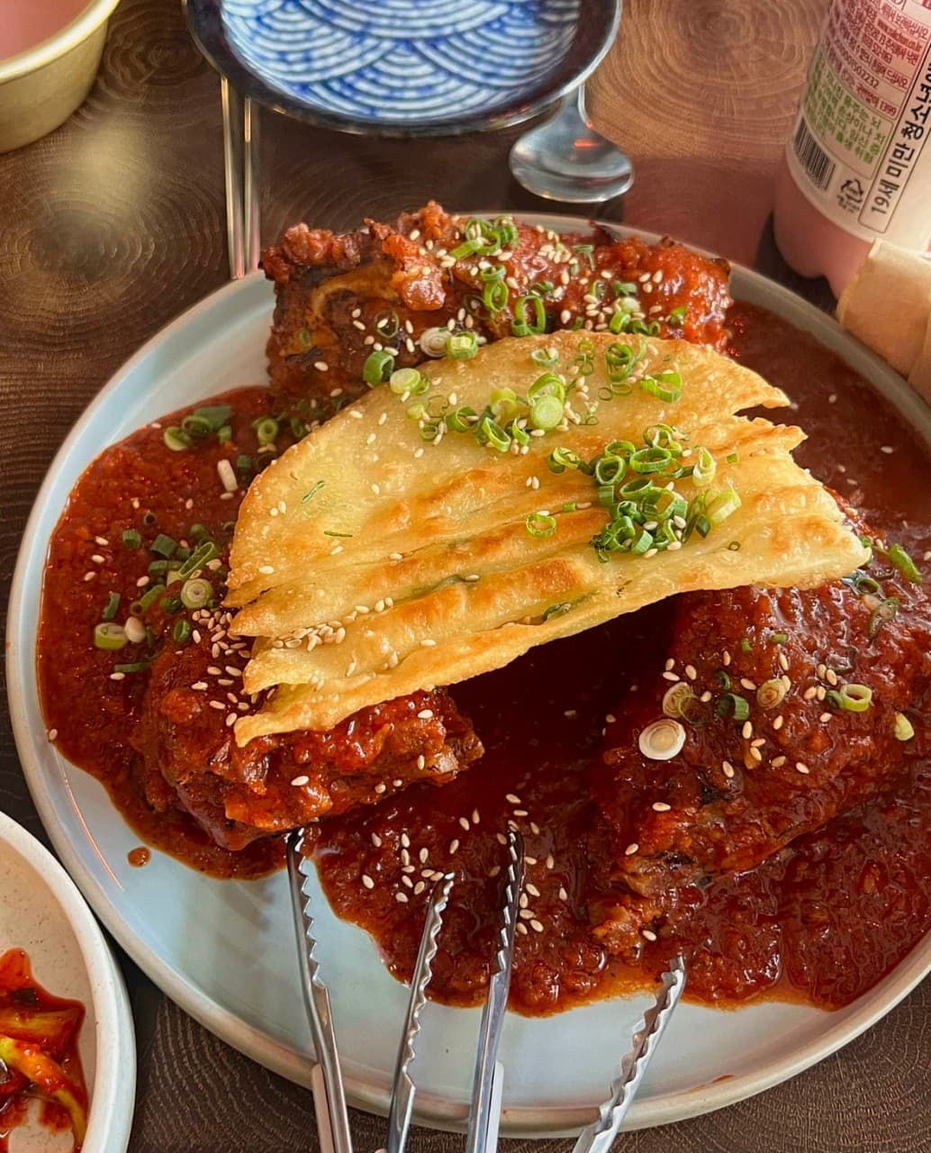 경기도 안양시 혼술 요리주점 이복실 경기 - 조명 아래 1인 다찌석 붉은 양념 고기와 바삭한 전. 파와 깨가 뿌려진 안주와 술병이 놓인 식탁 상황.