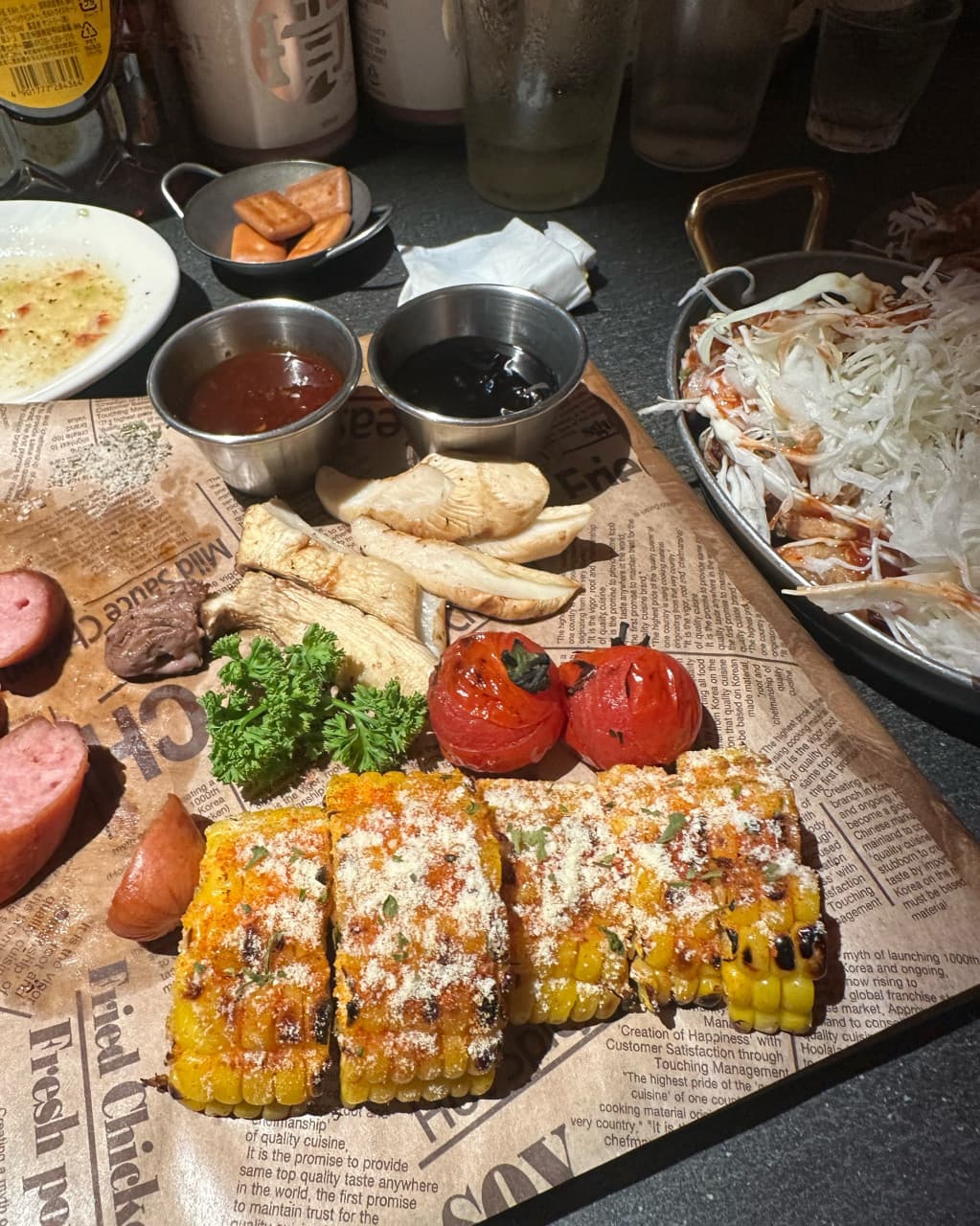 경기도 성남시 일편닭심 판교본점 경기 요리주점 다찌석 1인 혼술. 시즈닝된 옥수수 구이와 구운 채소가 종이 위에 차려진 질감과 어두운 조명 분위기를 나타냄.