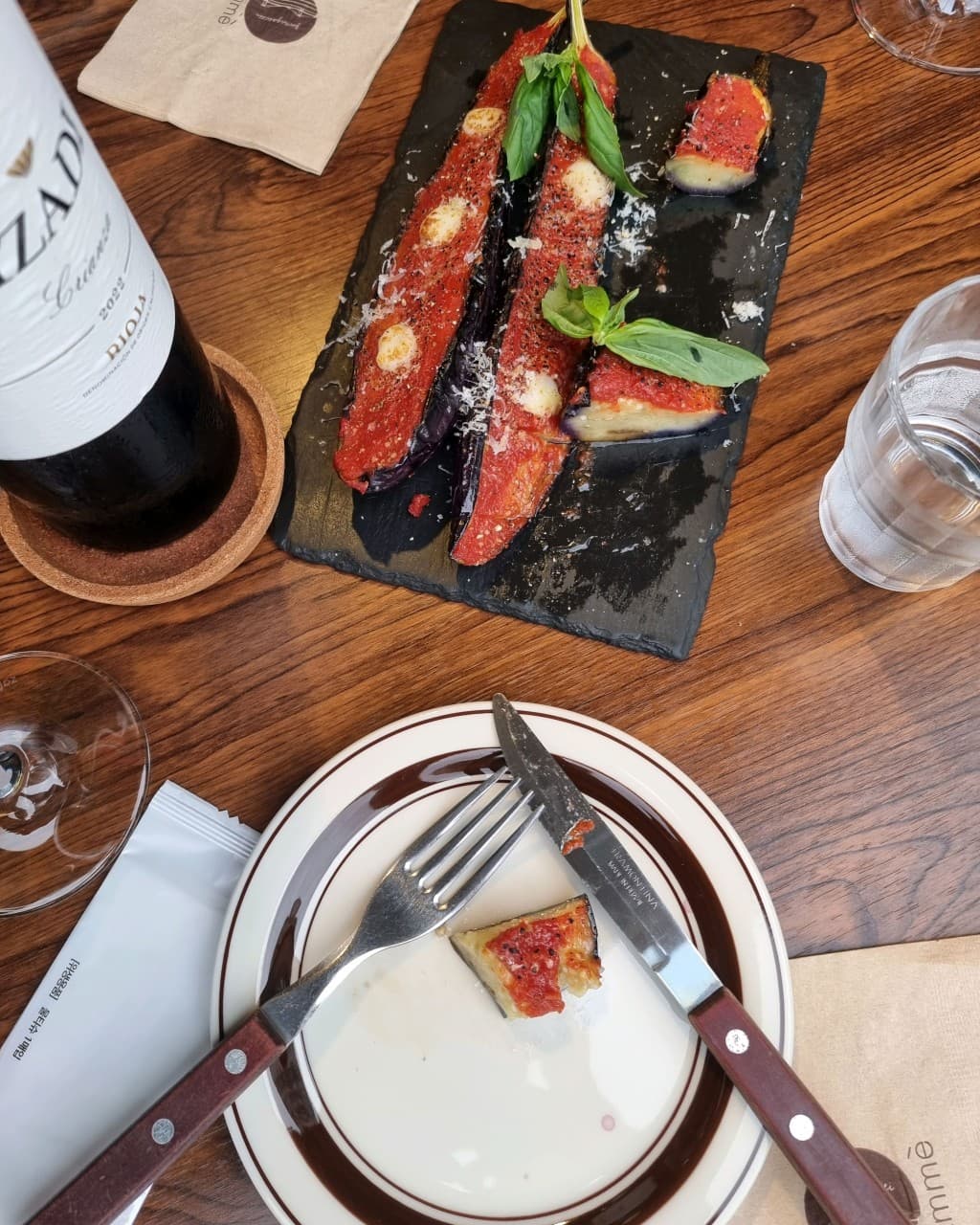 부산광역시 수영구 광안리남매 부산 와인 바 나무 테이블 위 붉은 소스와 바질을 곁들인 가지 요리. 1인 혼술 좌석에 놓인 레드 와인과 나이프, 포크가 놓인 접시.