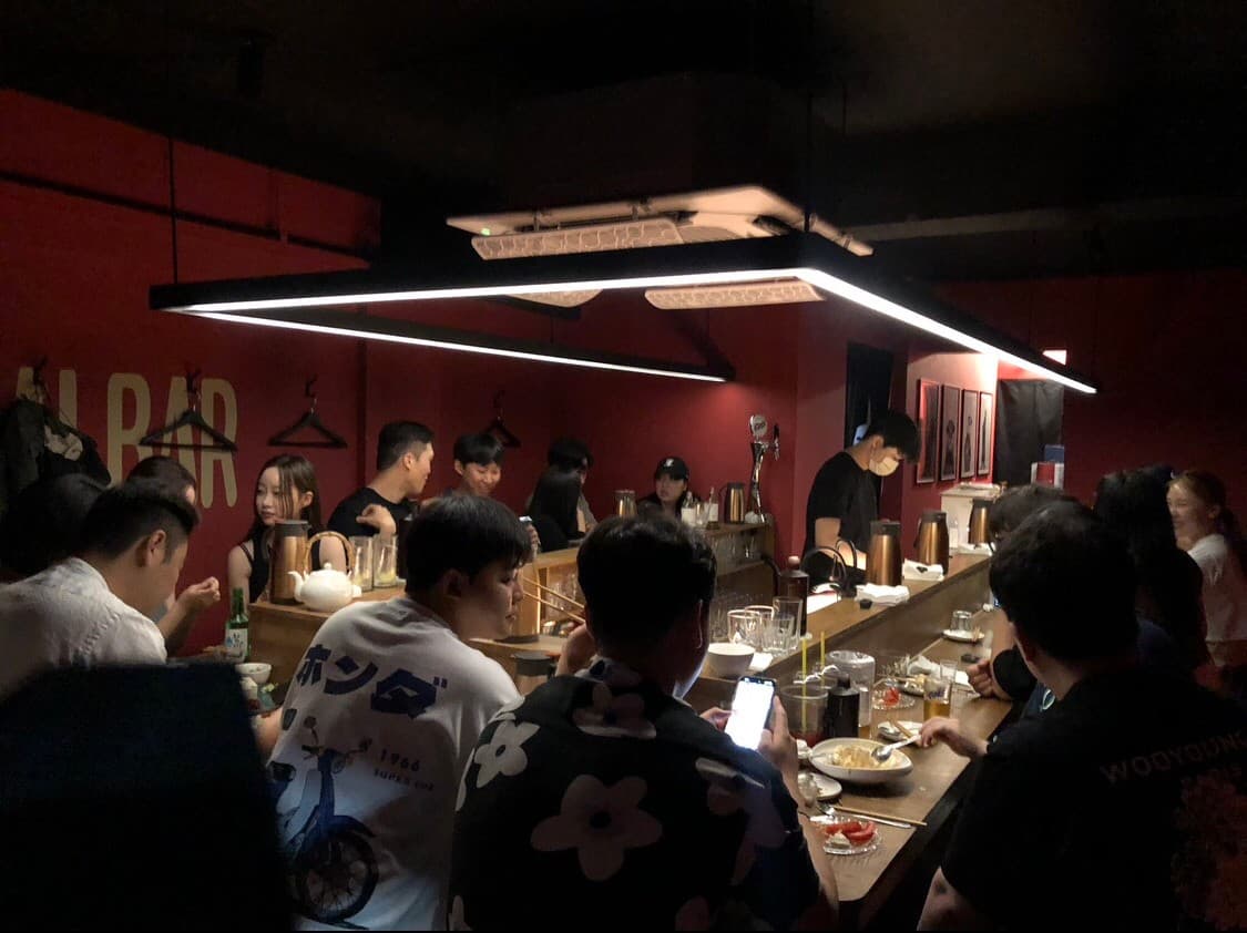 부산광역시 수영구 요리주점 수달바 광안 부산의 붉은 벽면과 사각형 조명 아래 다찌석. 1인 혼술이 가능한 목재 카운터 좌석에서 다양한 요리와 술을 곁들이는 상황.