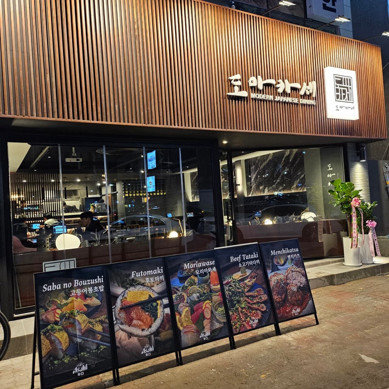 대구광역시 북구 이자카야 도마카세 칠곡3지구점 대구. 나무 간살 외벽 조명 아래 후토마끼와 사시미 메뉴판이 있고 통유리창 너머 내부 다찌석에서 1인 혼술이 가능한 공간 구성.