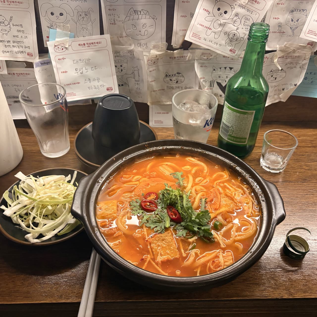 대구광역시 중구 이자카야미우 대구 이자카야의 나무 다찌석 1인 혼술 차림. 빨간 국물의 전골과 소주, 양배추 샐러드가 조명 아래 놓여 있고 벽에는 메모지가 가득하다.