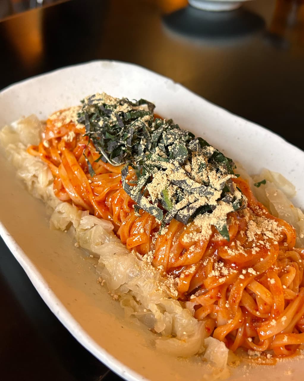 대구광역시 중구 도쿄테이스트 대구 이자카야 다찌석 조명 아래 놓인 1인 비빔면. 빨간 양념 국수 위에 김가루와 가루 고명이 뿌려진 음식과 백김치가 담긴 혼술 상차림.