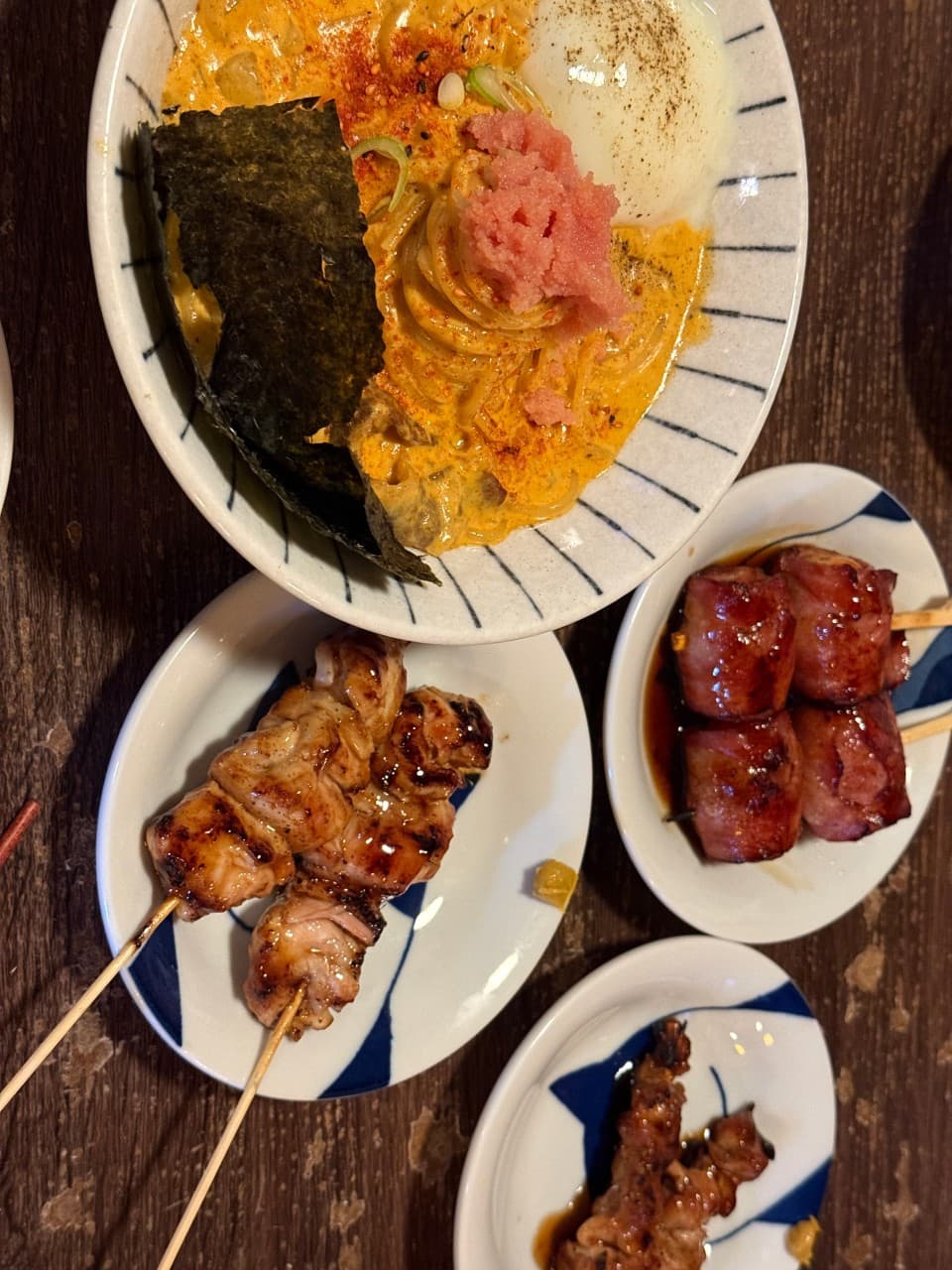 대구광역시 동구 이자카야 요시쿠 신서 본점 대구의 나무 테이블 위 명란 크림 파스타와 수란, 윤기 나는 닭꼬치가 차려진 1인 혼술 상황. 조명 아래 음식의 질감이 강조된 모습.