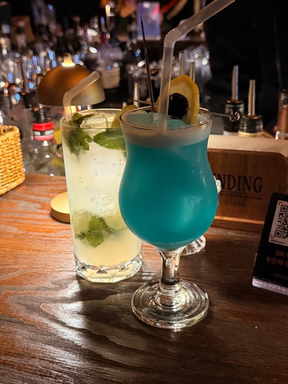 부산광역시 수영구 블렌딩바 부산혼술바 광안리점 이자카야 다찌석에 놓인 푸른색 칵테일과 라임 모히토. 노란 조명 아래 나무 질감의 테이블 위에서 1인 혼술을 즐기는 상황.