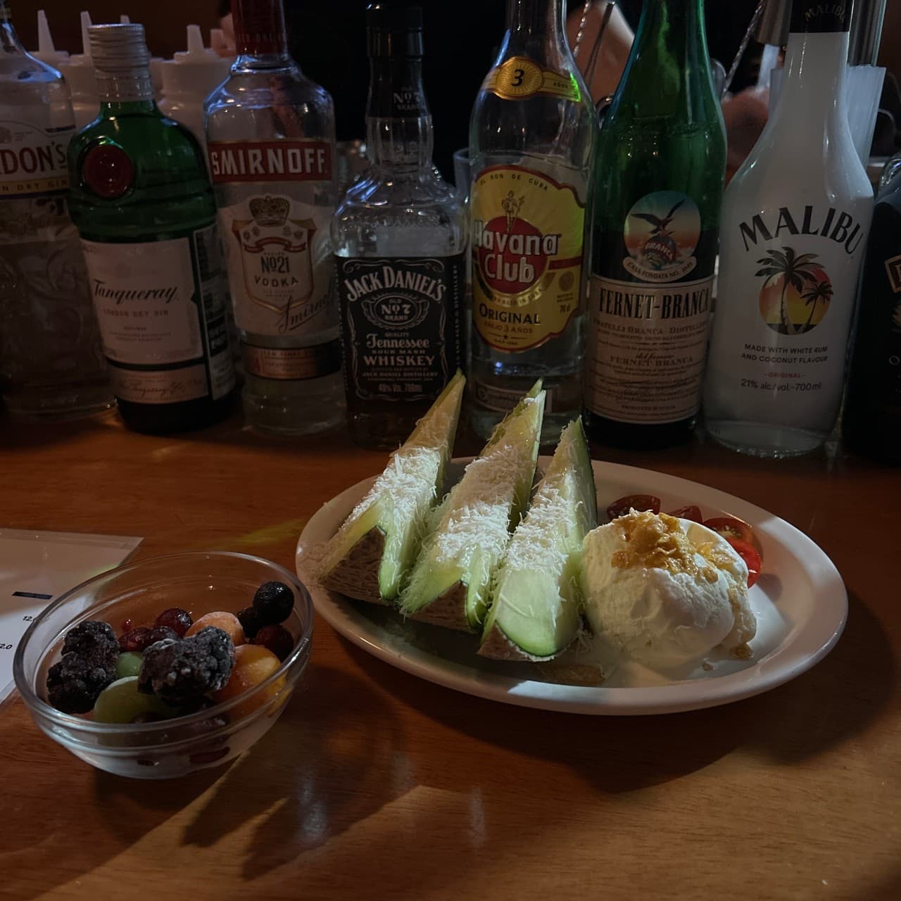 부산광역시 수영구 표류 부산 바(Bar) 다찌석에서 혼술하며 즐기는 치즈 멜론과 아이스크림. 1인 테이블 뒤로 늘어선 다양한 술병들과 노란 조명 아래 놓인 안주.