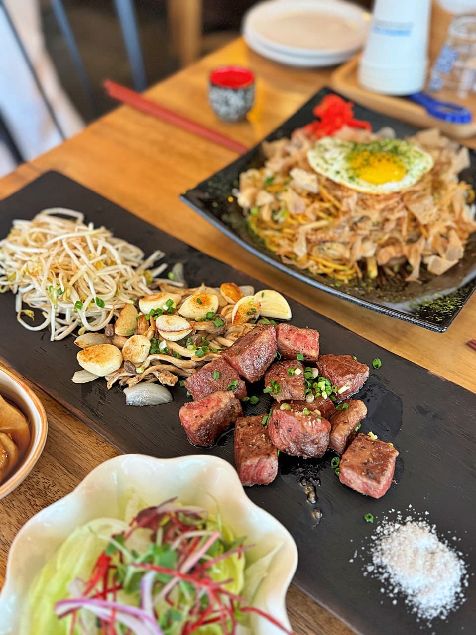 부산광역시 부산진구 요리주점 데판킹 부산 1인 다찌석 혼술. 조명 아래 육즙 가득한 큐브 스테이크와 구운 마늘, 계란 프라이를 올린 야키소바가 차려진 상차림.