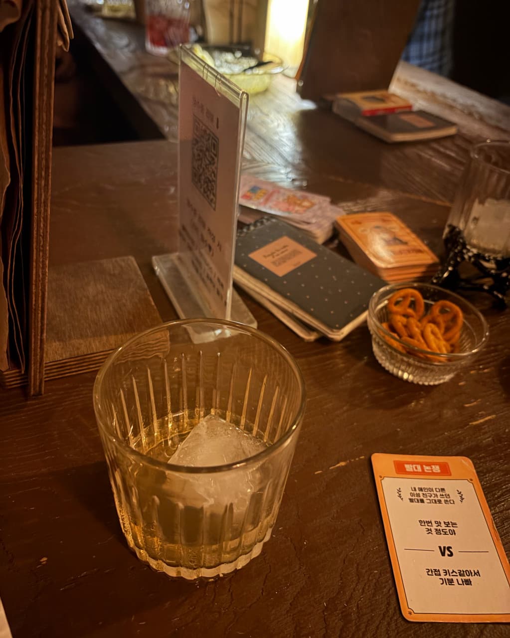 부산광역시 수영구 곁 부산혼술바 광안리점 바(Bar) 나무 다찌석에 놓인 커다란 얼음 잔과 프레첼. 질문 카드와 함께 1인 혼술을 즐기는 낮은 조도의 실내 상황.