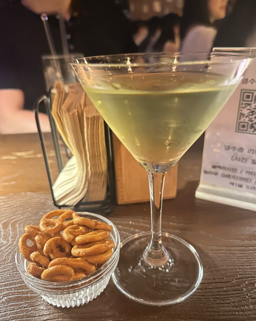 부산광역시 수영구 곁 부산혼술바 광안리점 바(Bar)의 조명 아래 나무 테이블 다찌석에 차려진 칵테일과 프레첼. 혼술을 즐기는 1인석 자리 구성.