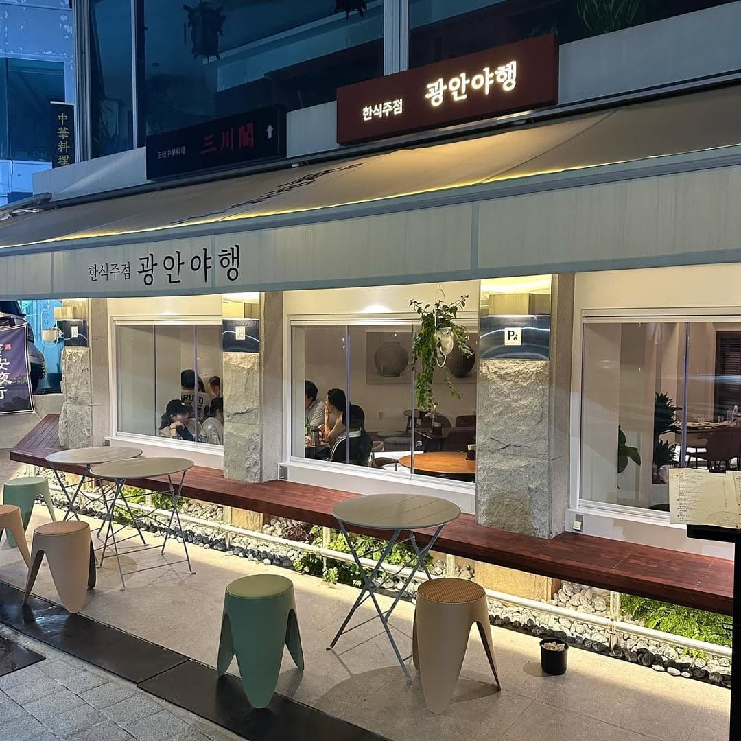 부산광역시 수영구 광안야행 부산 요리주점. 조명 아래 야외 원형 테이블과 1인 혼술 가능한 창가 좌석이 배치된 석재 기둥과 식물이 어우러진 외부 전경.