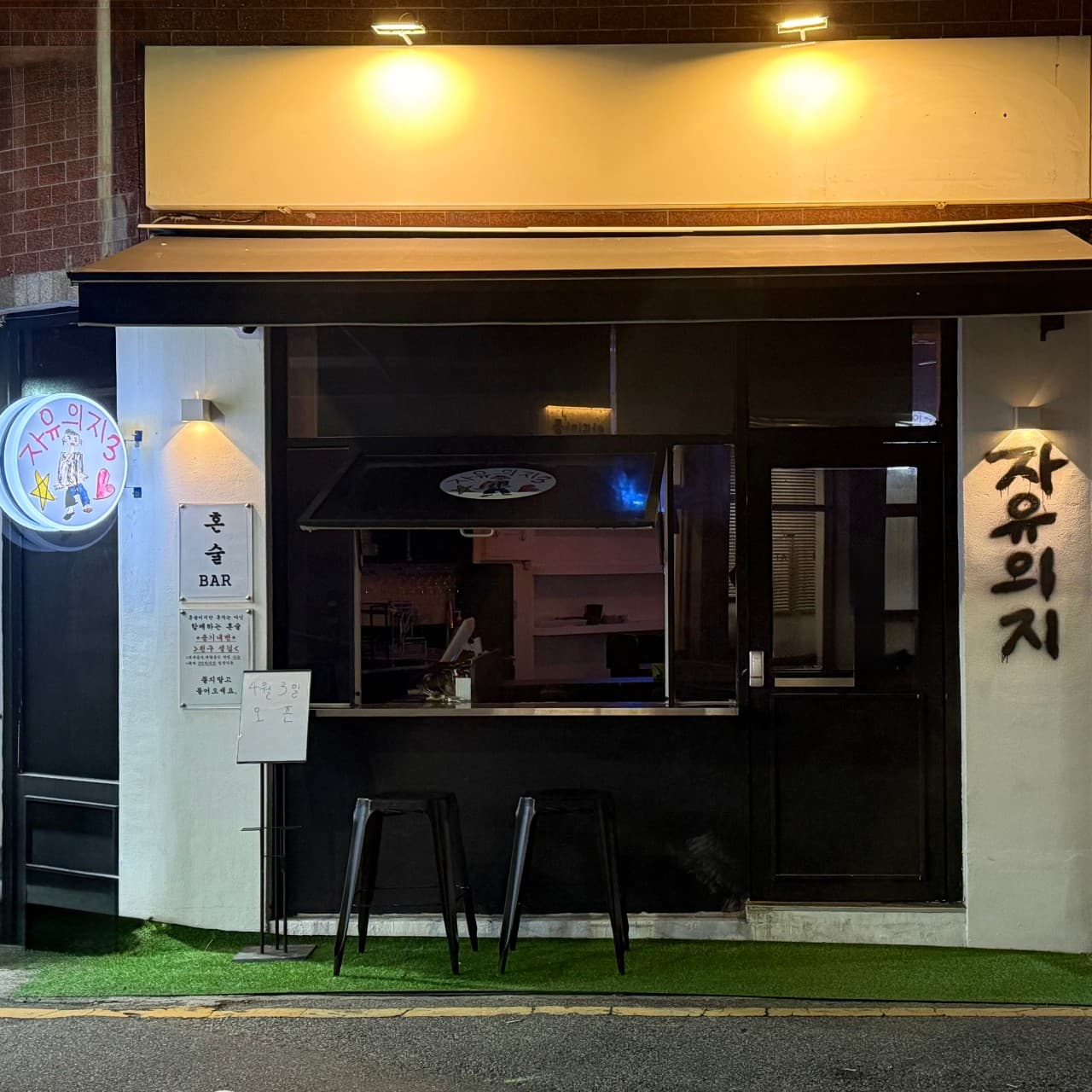 부산광역시 수영구 자유의지 혼술바 부산광안리점 바(Bar) 외부 전경. 노란 조명이 비치는 흰색 벽면에 1인 다찌석 의자와 혼술 전용 간판이 배치된 모습.