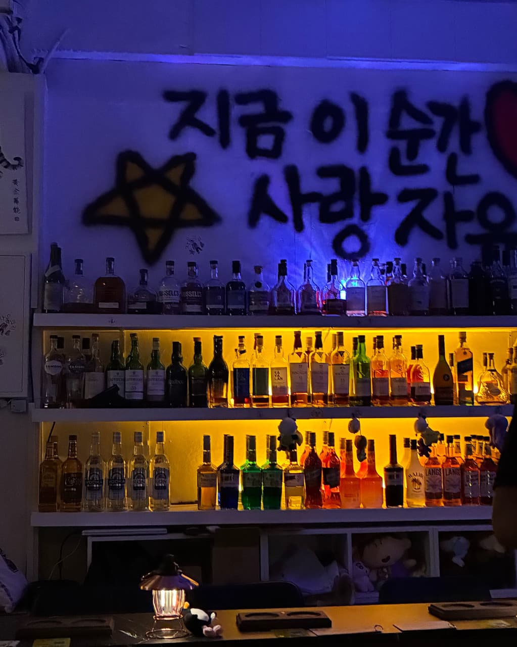 부산광역시 수영구 자유의지 혼술바 부산광안리점의 노란 조명 아래 진열된 다양한 주류와 1인 다찌석. 벽면 낙서와 등불이 조화를 이루는 혼술 바(Bar) 실내 전경
