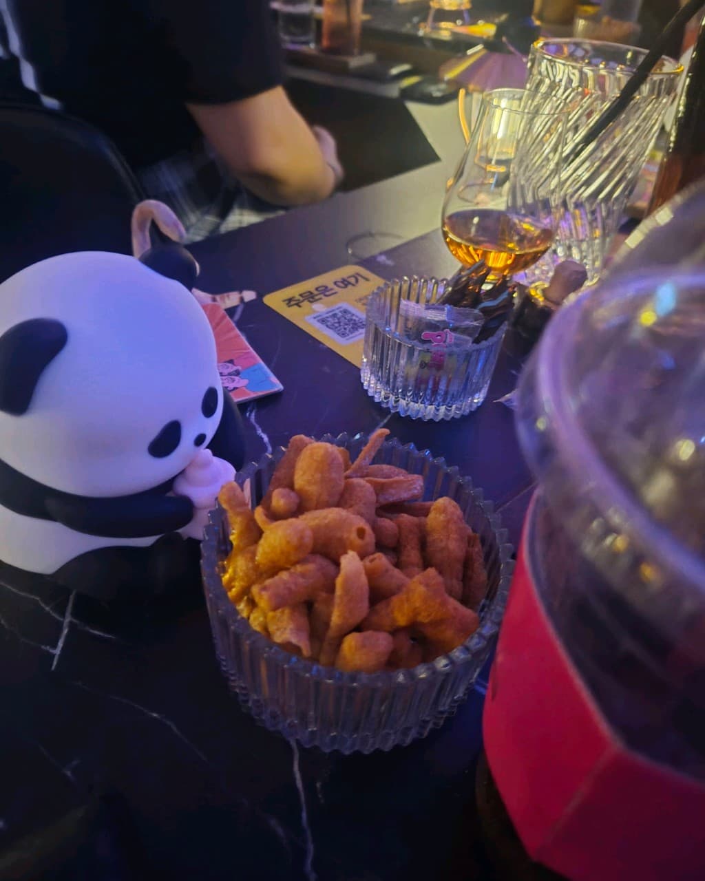 부산광역시 수영구 자유의지 혼술바 부산광안리점 바(Bar) 1인 다찌석에 차려진 위스키와 바삭한 질감의 과자. 판다 피규어와 QR 코드가 놓인 어두운 조명 아래의 혼술 상황.
