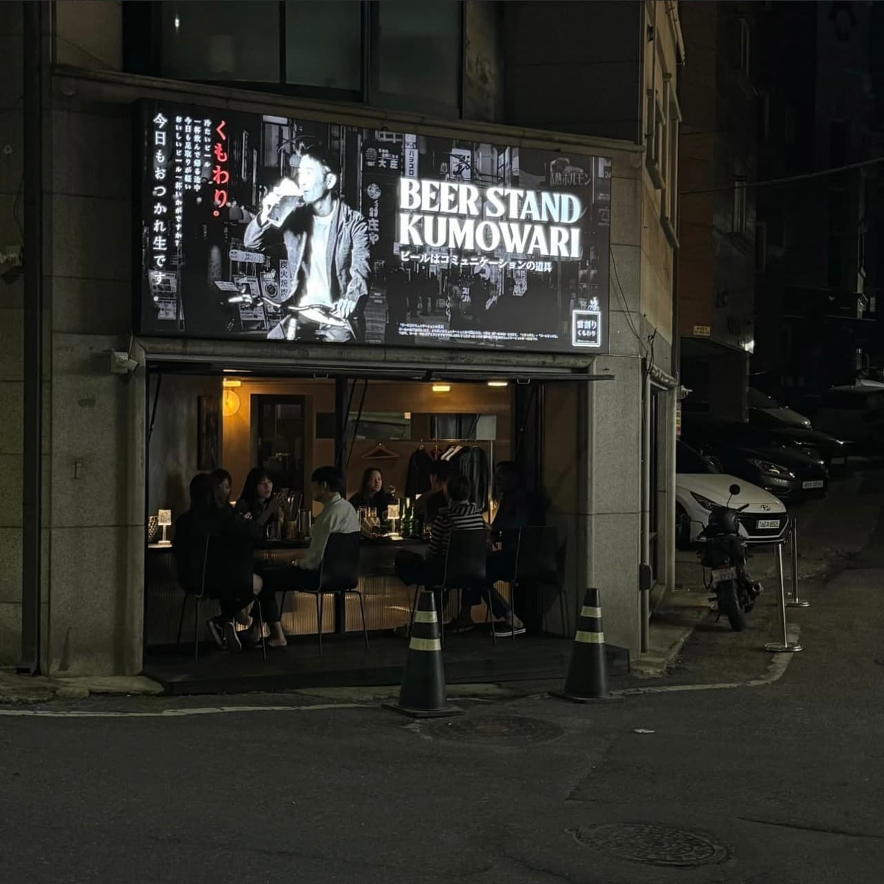 대전광역시 서구 이자카야 비어스탠드쿠모와리 대전. 밤거리 환한 간판 아래 개방된 다찌석에서 맥주를 즐기는 상황. 1인 혼술을 위한 일자형 카운터석과 노란색 조명이 비치는 매장 내부.