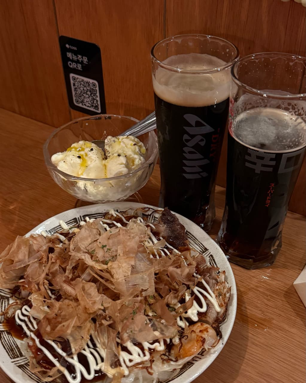대전광역시 유성구 맥주 호프 타치노미 럽밤 대전 - 황색 조명 아래 나무 다찌석에서 즐기는 흑맥주와 가쓰오부시가 올라간 요리가 차려진 1인 혼술 상황
