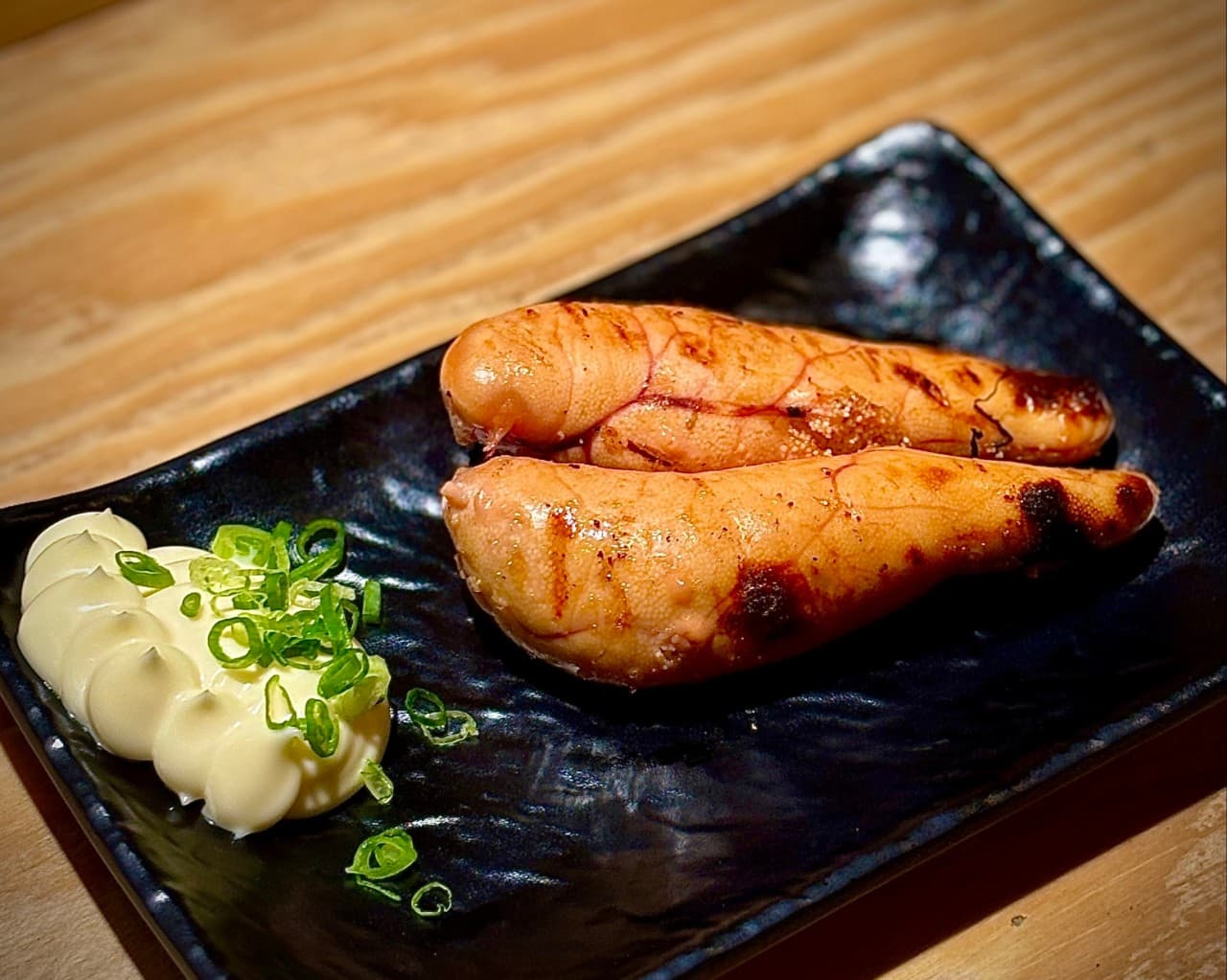 대전광역시 서구 혼술 이자카야 토리아에즈 대전둔산점 - 다찌석 나무 테이블 위 조명을 받은 명란구이. 검은 접시에 노릇하게 구워진 명란과 마요네즈, 쪽파가 차려진 1인 메뉴 구성.