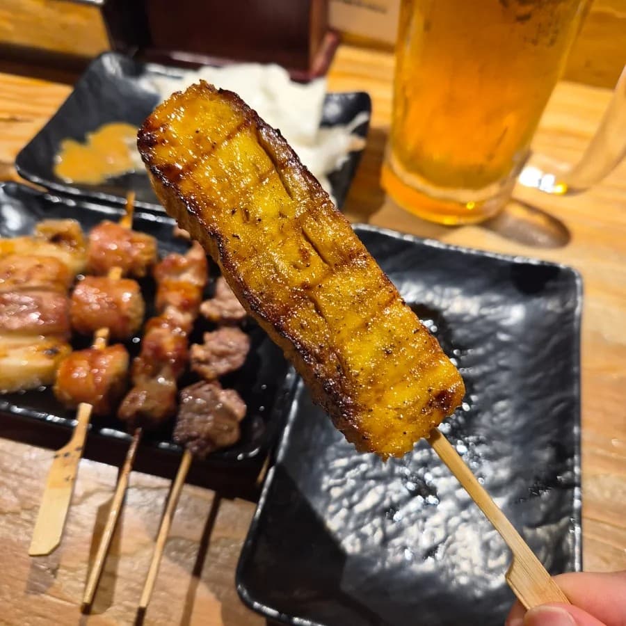 대전광역시 서구 토리아에즈 대전둔산점 이자카야 다찌석. 노릇하게 구워진 꼬치구이의 질감과 생맥주가 차려진 1인 혼술 상황. 나무 테이블 위로 밝은 조명이 비치는 구성.