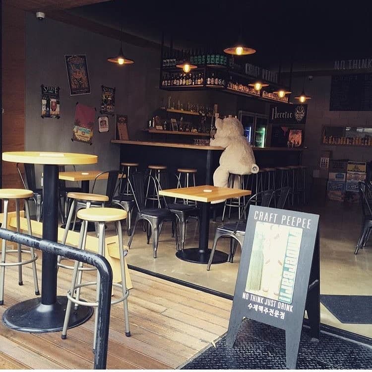 대전광역시 서구 크래프트피피 대전 맥주 호프 매장. 전구 조명 아래 바 좌석에 곰인형이 홀로 앉아 혼술하는 상황. 나무 질감의 테이블과 철제 의자가 배치된 개방형 공간.