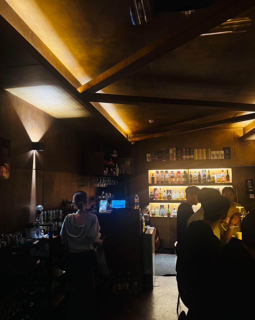 인천광역시 남동구 혼술바 고도 구월점 인천 바(Bar) - 노란 조명 아래 다찌석에 앉아 혼술을 하는 사람들과 다양한 주류 병이 진열된 선반이 있는 내부 공간