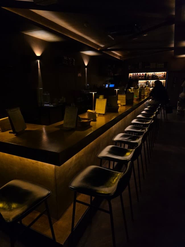 인천광역시 남동구 혼술바 고도 구월점 인천 바(Bar) 내부의 어두운 조명과 긴 다찌석. 일렬로 배치된 1인석 의자와 메뉴판이 놓인 카운터 테이블.