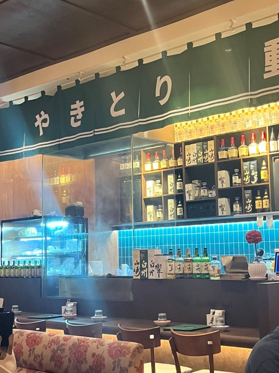 인천광역시 남동구 야키토리동작 인천시청점 이자카야의 1인 다찌석과 위스키 진열대. 초록색 노렌 아래 간접 조명이 비치는 카운터 좌석에서 혼술을 즐기는 상황을 묘사함.