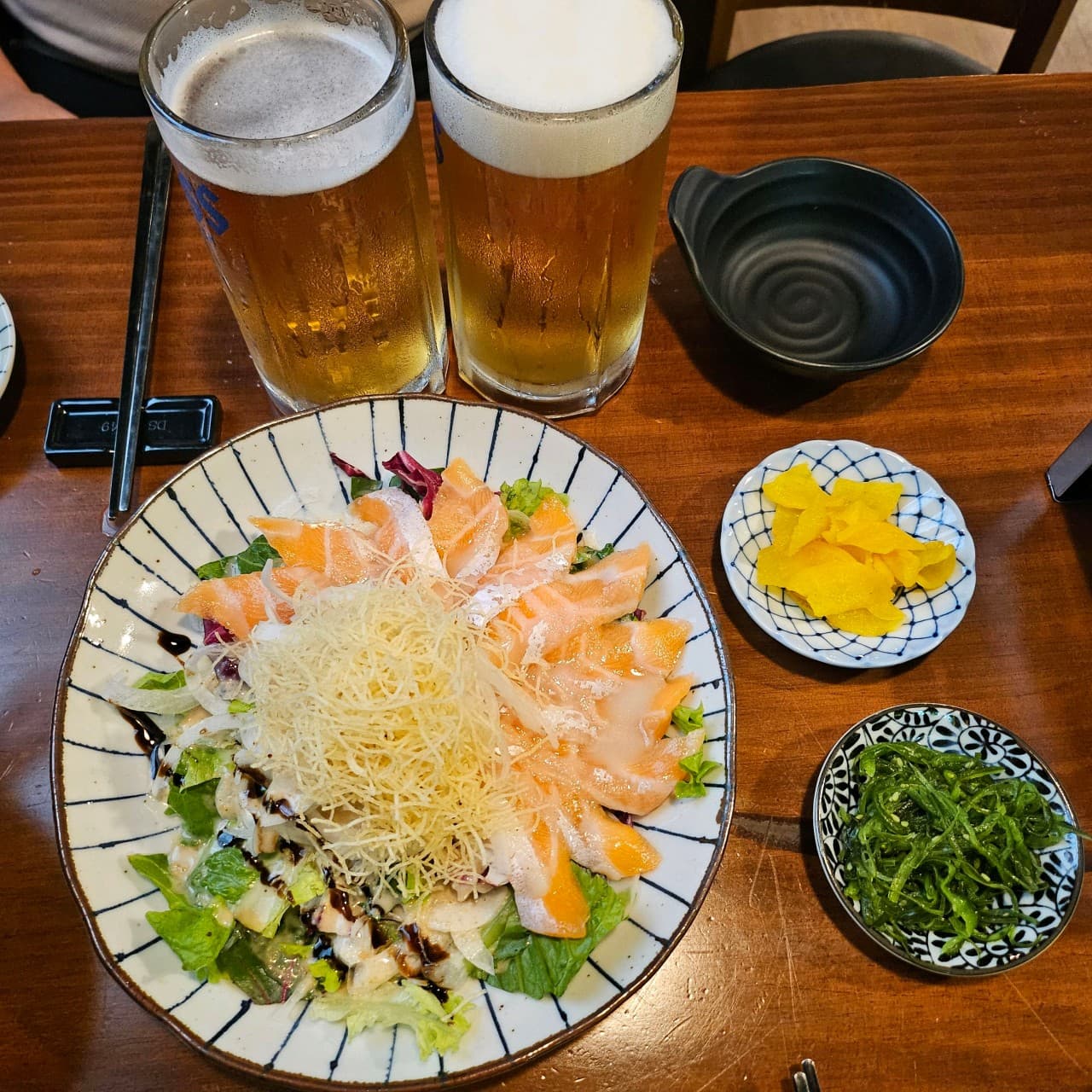 인천광역시 서해구 니치니치 인천 이자카야 나무 테이블 위 생맥주 두 잔과 연어 샐러드. 1인 다찌석에서 즐기는 혼술 차림과 가지런한 밑반찬 구성.