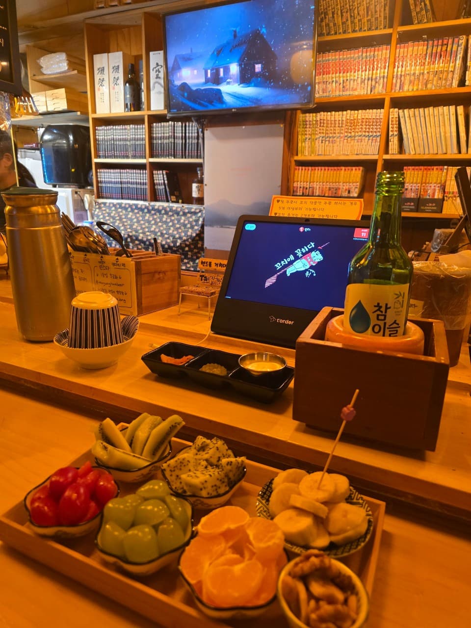 인천광역시 부평구 무궁화꼬치피었습니다 인천 다찌석 혼술. 주황빛 조명 아래 과일 안주와 소주가 차려진 기타 선술집 카운터석과 만화책이 가득한 배경.