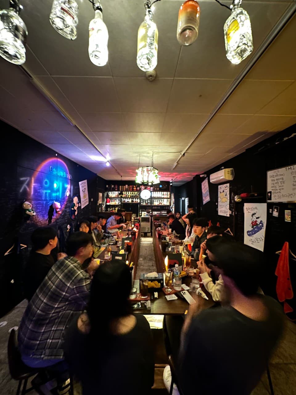 제주특별자치도 제주시 자유의지 혼술바 제주본점 바(Bar) 내부의 병 모양 조명 아래 긴 다찌석 좌석 배치. 네온 사인과 1인 손님들이 혼술을 즐기는 카운터석 상황을 보여줌.