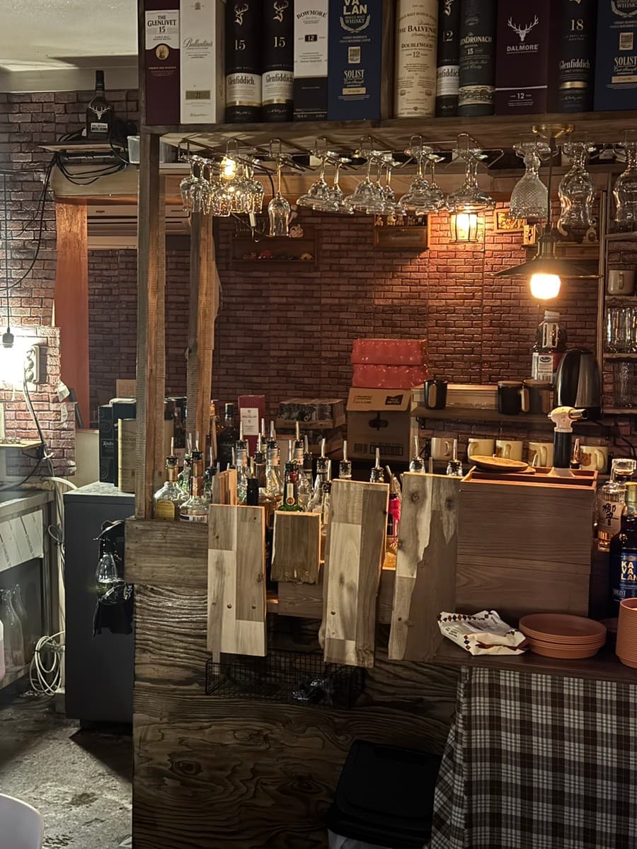 제주특별자치도 제주시 곁 제주혼술바 탑동점 본점 바(Bar) 내부. 벽돌 벽과 따뜻한 색감의 조명 아래 우드 다찌석과 다양한 위스키병, 매달린 와인잔이 조화를 이루는 1인 혼술 공간.