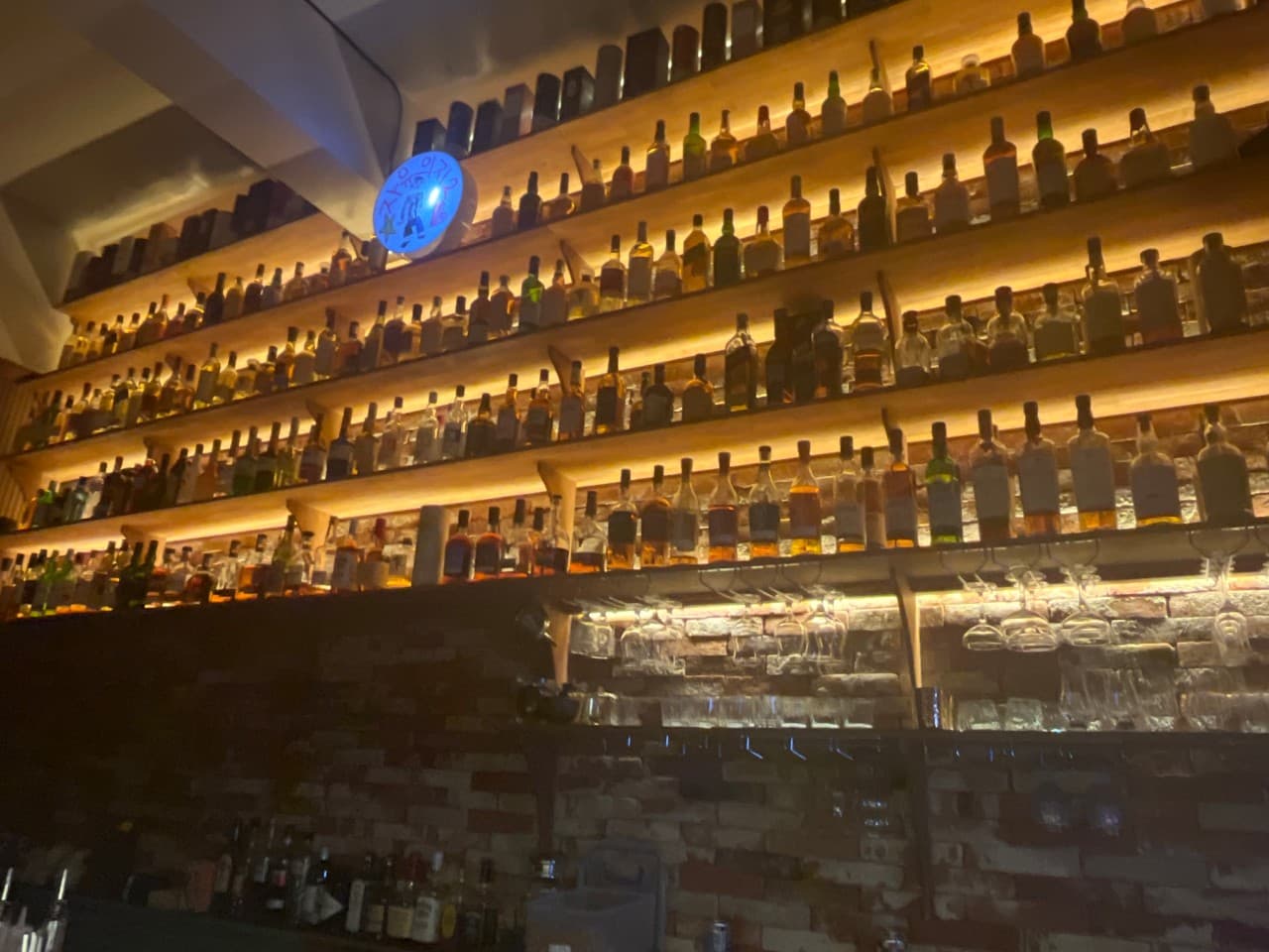 제주특별자치도 서귀포시 자유의지 혼술바 제주서귀포점 바(Bar)의 주황색 조명이 비치는 목재 선반과 수많은 술병들. 벽돌 벽면에 걸린 와인잔과 혼술을 위한 다찌석 배치를 보여줌.
