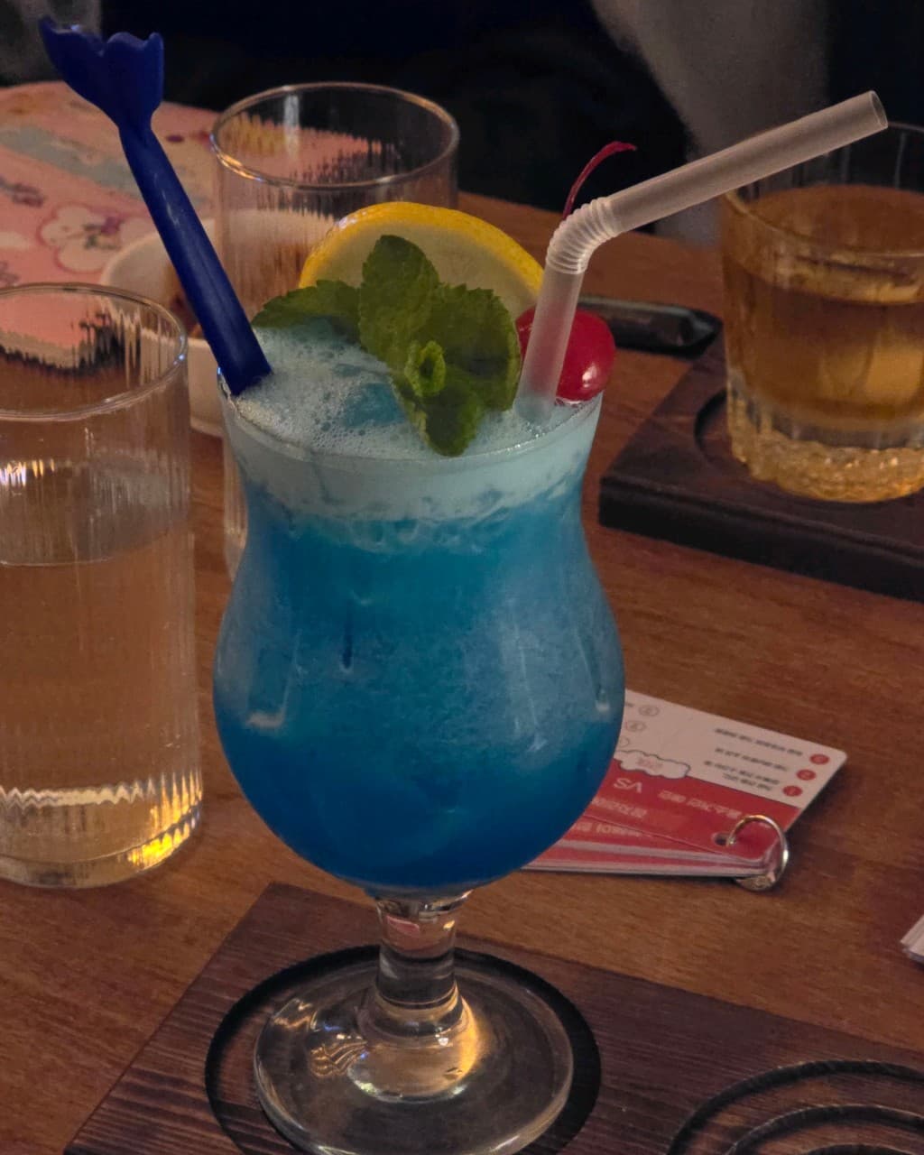제주특별자치도 서귀포시 자유의지 혼술바 제주서귀포점 바(Bar)의 나무 테이블 위 레몬과 민트가 장식된 파란색 칵테일. 조명 아래 놓인 1인용 술잔과 메뉴판이 있는 혼술 상황.