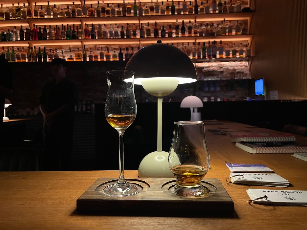 제주특별자치도 서귀포시 자유의지 혼술바 제주서귀포점 바(Bar) 다찌석 조명 아래 놓인 위스키. 벽면 술병 진열장과 나무 테이블에서 즐기는 1인 혼술 상황