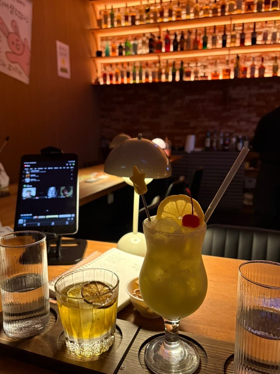 제주특별자치도 서귀포시 자유의지 혼술바 제주서귀포점 바(Bar)의 1인석. 레몬 칵테일과 태블릿이 놓인 다찌석 너머로 조명을 받은 수많은 술병이 진열됨.