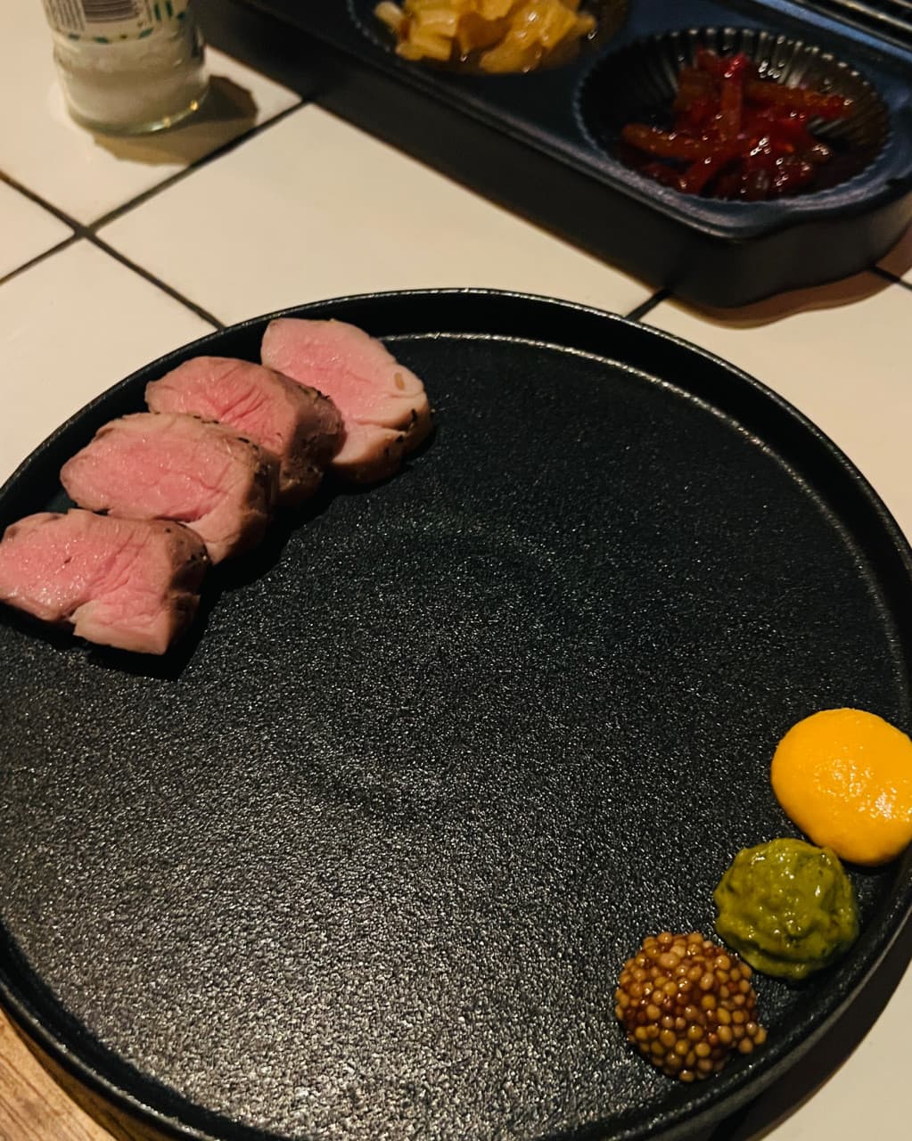 제주특별자치도 서귀포시 명단 BBQ BAR 제주 1인 다찌석 조명 아래 선분홍색 육질의 고기와 세 가지 소스. 기타 다이닝 바의 타일 테이블 위에 배치된 구성.