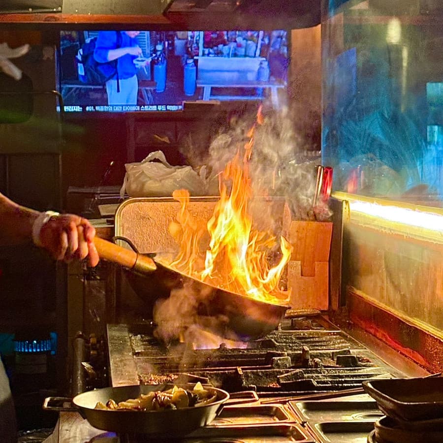 대전광역시 중구 심야식당11월6일 대전의 다찌석 앞 주방에서 강한 불길로 웍질하는 상황. 어두운 조명 속 혼술을 즐기며 기타 안주가 조리되는 과정을 직관하는 시점.