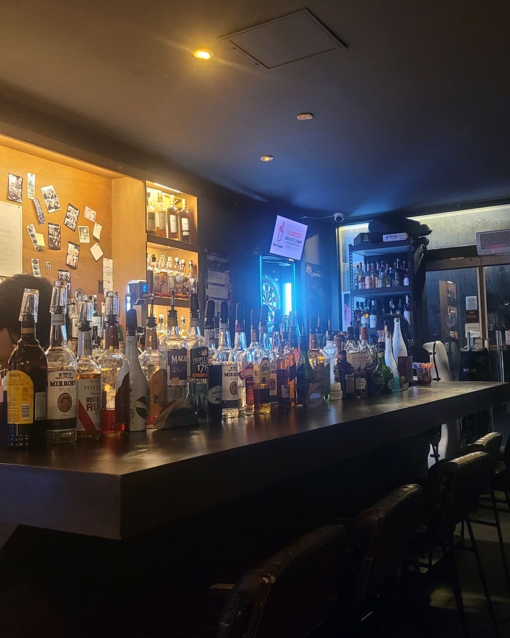 대전광역시 동구 공강 대전 바(Bar) 다찌석에 진열된 다양한 위스키 병들과 노란 상단 조명. 1인 방문이 가능한 어두운 실내 카운터 좌석과 배경의 다트 기계로 구성된 혼술 공간.