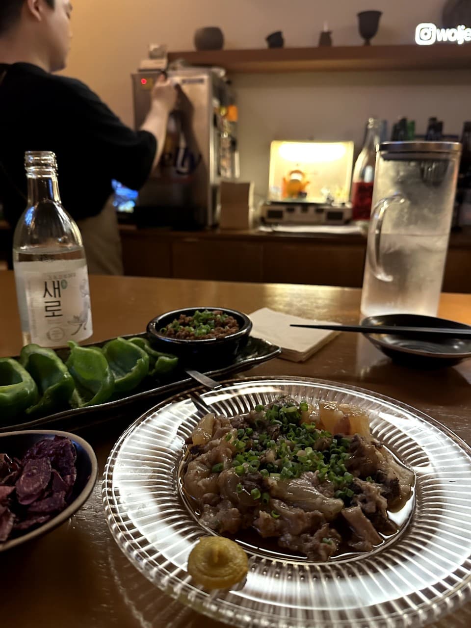 대전광역시 서구 월정 대전 이자카야 다찌석에 차려진 고기 요리와 소주 한 병. 노란 조명 아래 1인 테이블에 놓인 피망과 안주가 혼술 상황을 보여줌.