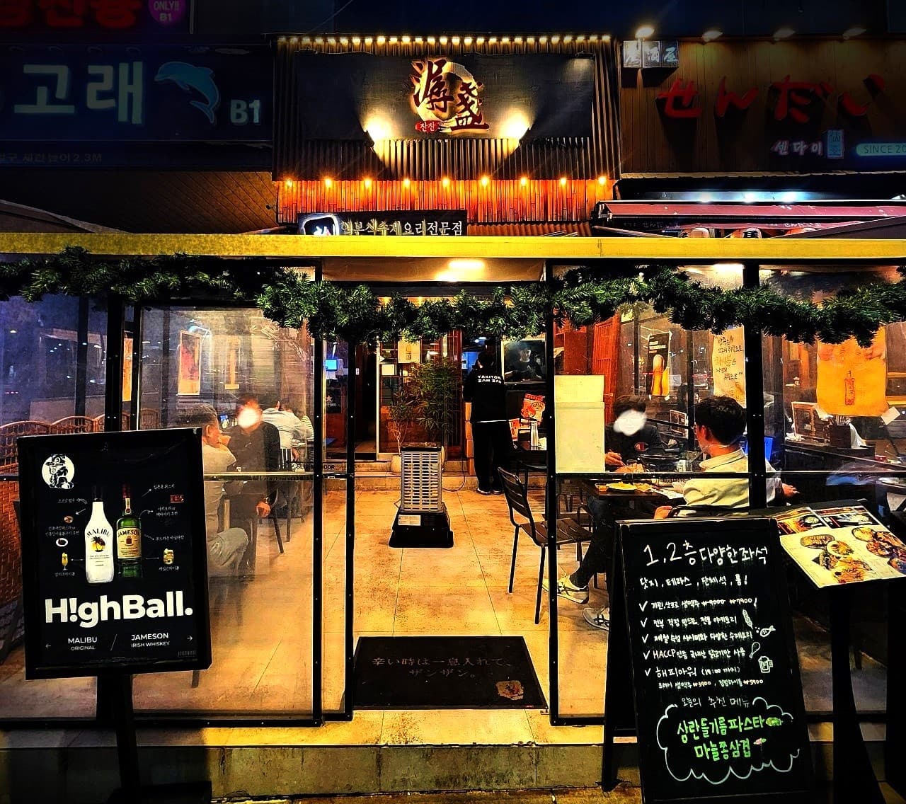 경기도 성남시 혼술 이자카야 잔잔 정자역점 경기 - 전구 조명 아래 1인 다찌석과 테라스 좌석, 하이볼 메뉴판이 배치된 유리창 너머의 내부 전경