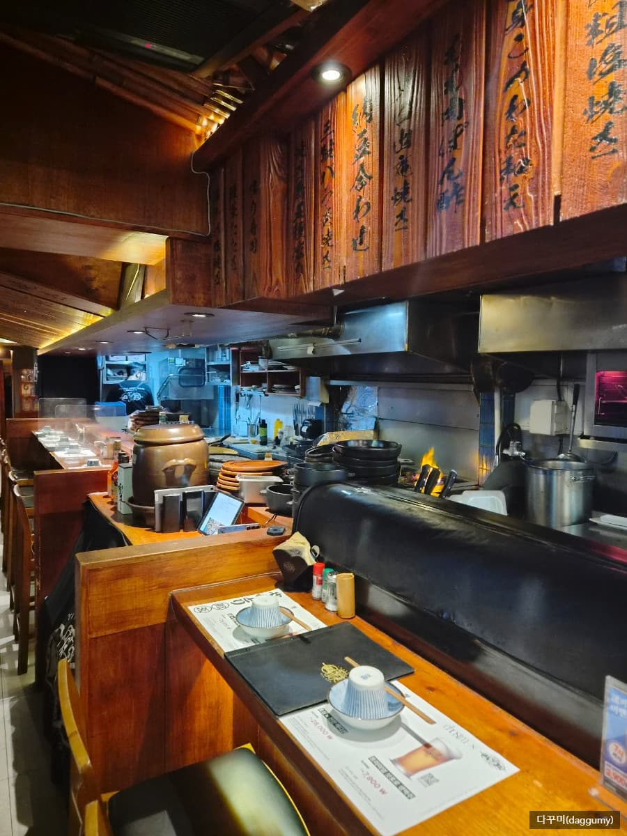 경기도 성남시 잔잔 정자역점 경기 이자카야의 다찌석. 주황색 조명 아래 1인용 식기가 세팅된 테이블과 불길이 보이는 오픈 주방이 어우러진 혼술 공간.