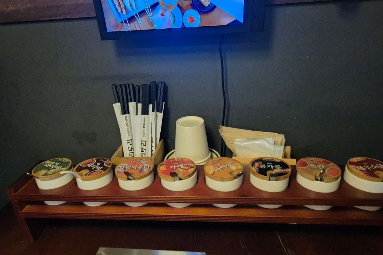 경기도 수원시 도리토리 수원곡반정점 경기 요리주점 다찌석에 비치된 8가지 양념통과 1인용 식기류. 혼술 상황에 맞춰 조명 아래 소스 선반과 젓가락이 정렬된 모습.