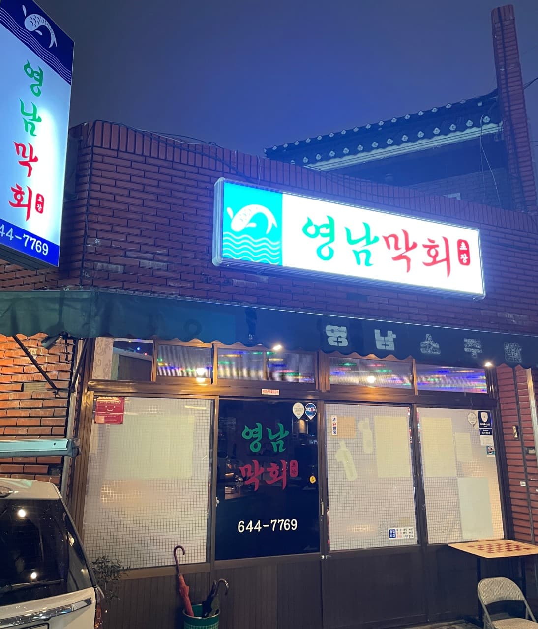 대구광역시 달서구 영남막회초장 상인본점 대구. 적벽돌 질감의 외벽 위 밝은 간판과 1인 혼술용 외부 좌석이 놓인 기타 식당 입구의 야간 조명.