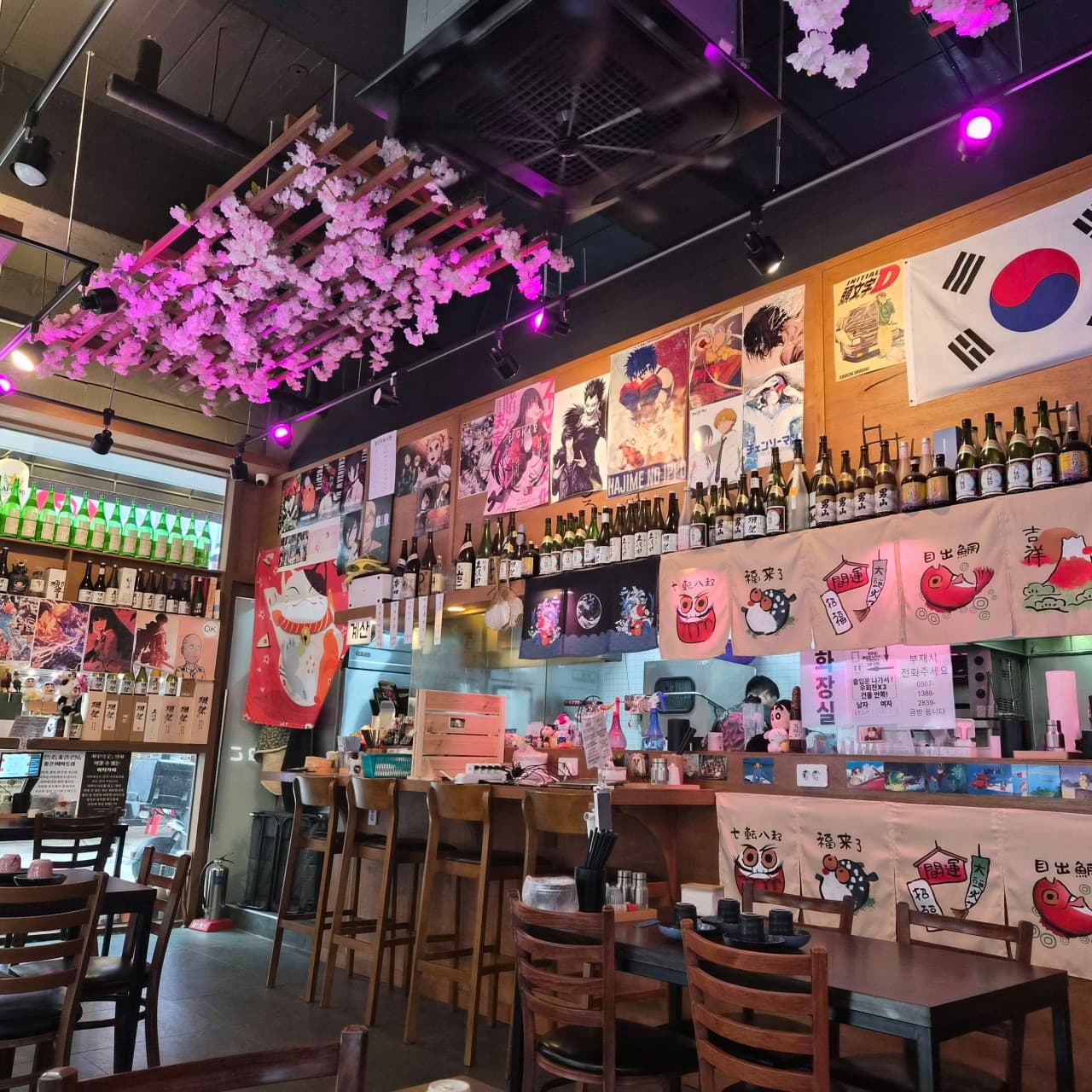 인천광역시 영종구 시마네코 인천 이자카야의 보라색 조명과 벚꽃 장식. 벽면의 애니메이션 포스터와 사케 병들 아래로 혼술용 다찌석과 1인 좌석이 배치된 실내 전경.