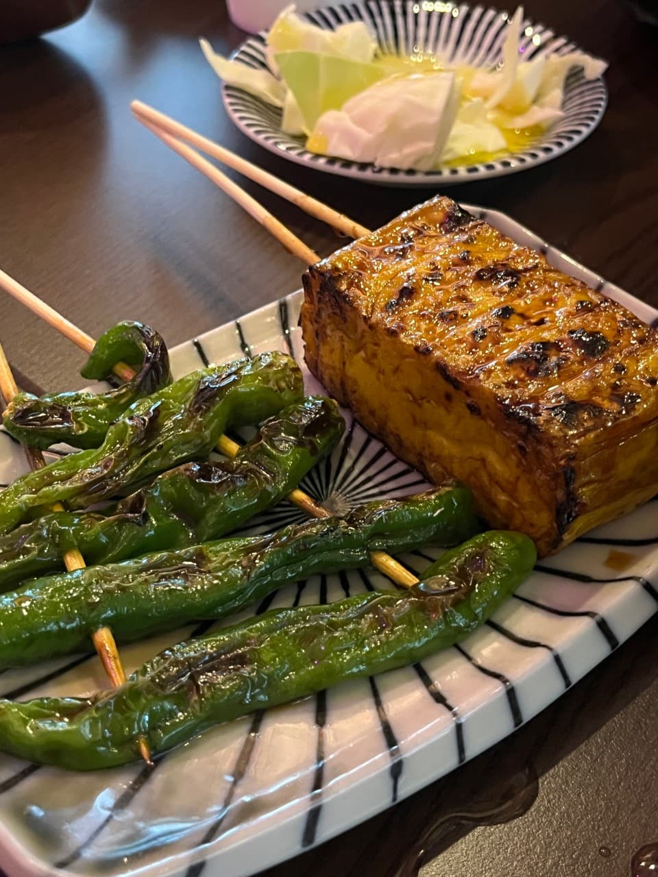 인천광역시 영종구 시마네코 인천 이자카야 다찌석 조명 아래 놓인 1인 꽈리고추와 두부 꼬치구이. 혼술 상황의 윤기 있는 안주 질감과 양배추가 차려진 테이블.