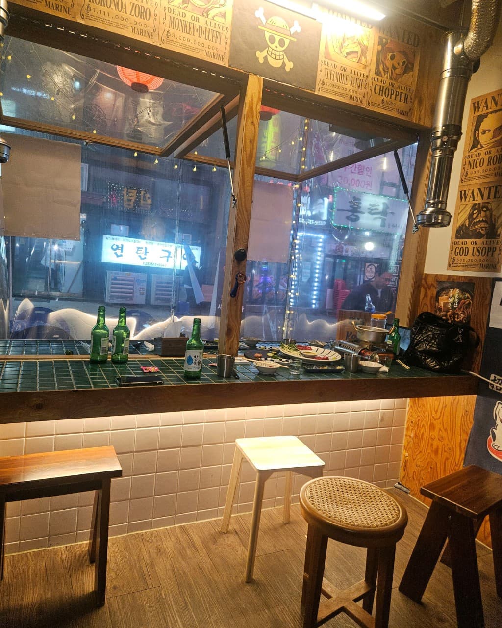 인천광역시 남동구 이자카야 육회천지 인천시청점의 타일 카운터 다찌석. 창가 1인 혼술 자리에 소주병과 안주 접시가 놓인 상황. 벽면의 캐릭터 포스터와 하단 간접 조명이 비치는 목재 의자.