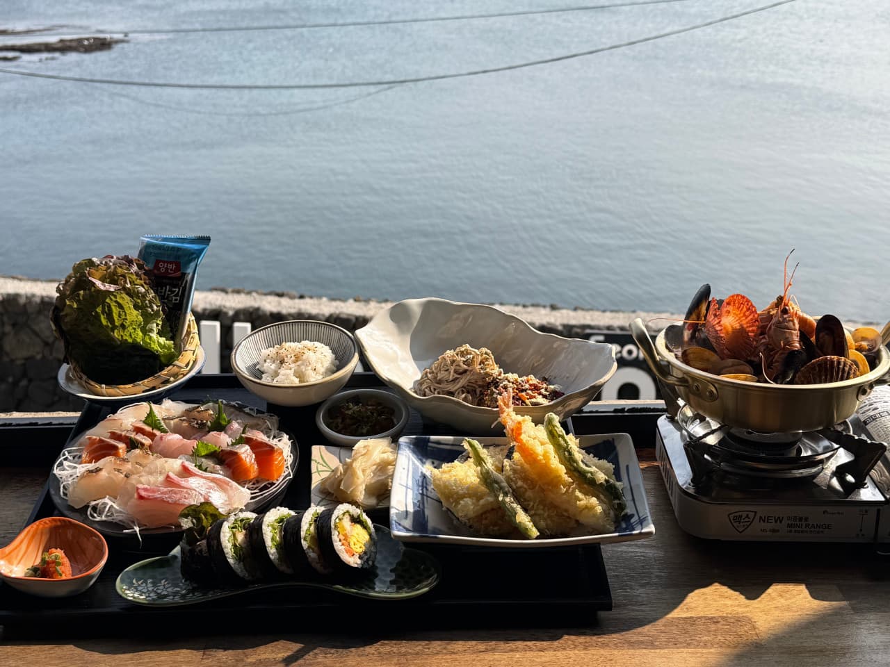 제주특별자치도 서귀포시 통발 성산점 제주 바다 전망 다찌석에 놓인 1인 해산물 정식. 햇살 아래 사시미와 해물탕, 튀김 등 기타 메뉴가 차려진 혼술 상차림.