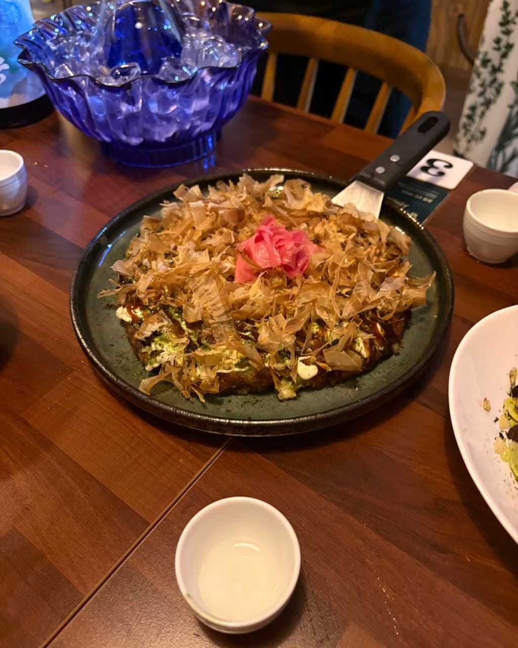 울산광역시 남구 혼술 이자카야 고꼬 - 조명 아래 나무 테이블 위 가쓰오부시가 덮인 오코노미야키와 사케 잔이 놓인 1인 좌석 상황