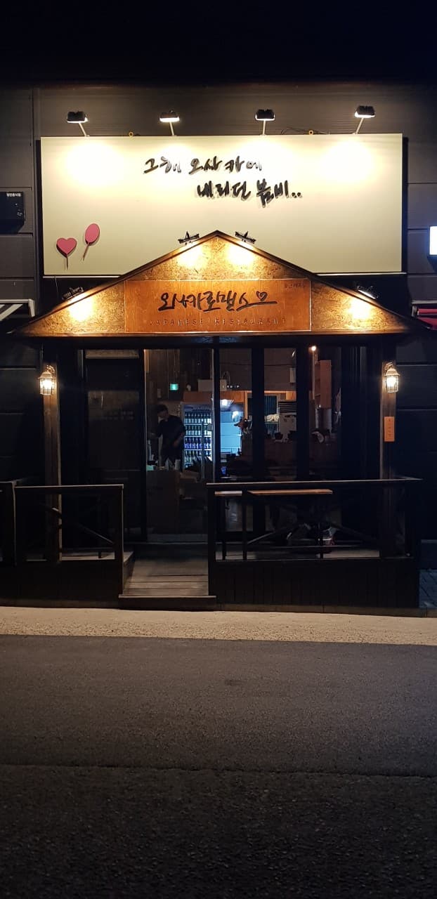 울산광역시 북구 오사카로맨스 매곡점 이자카야. 노란 조명이 비추는 나무 소재 입구와 다찌석이 보이는 유리창 너머로 1인 혼술 분위기가 전해짐.
